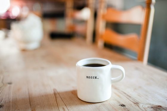 A white coffee mug with coffee in it. It has the word "BEGIN" on it, and sits on a wooden table.