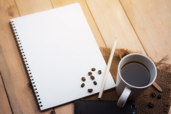 Mug of coffee on a square of burlap. A journal and pencil lay next to it. There are coffee beans scattered around the cup and on top of the journal.
