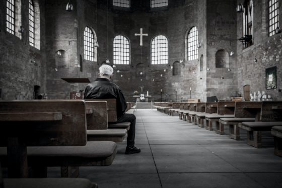 white man with gray hair faces away from camera, sitting in empty stone church.