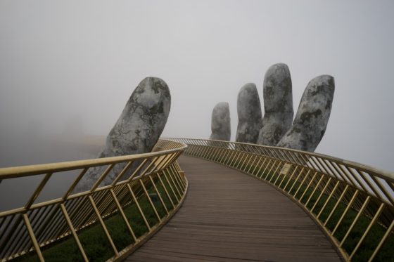 A footbridge in the fog. The bridge is held up by a concrete hand.