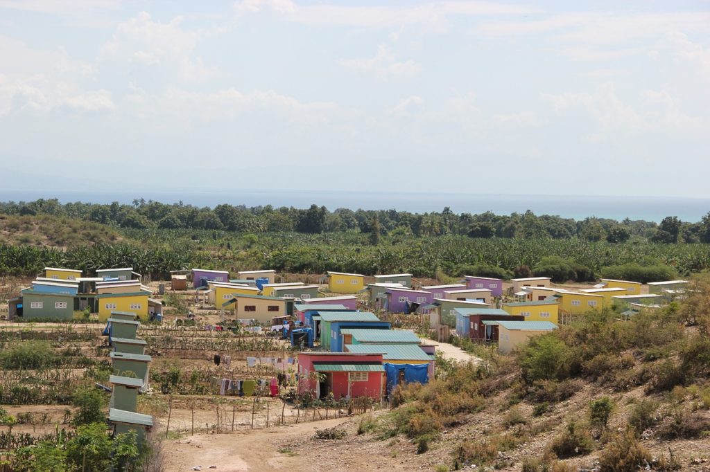 A view of a small village in Haiti. There are a dozen small cinderblock houses painted in pastel colors, some with private gardens behind them growing corn or tomatoes.