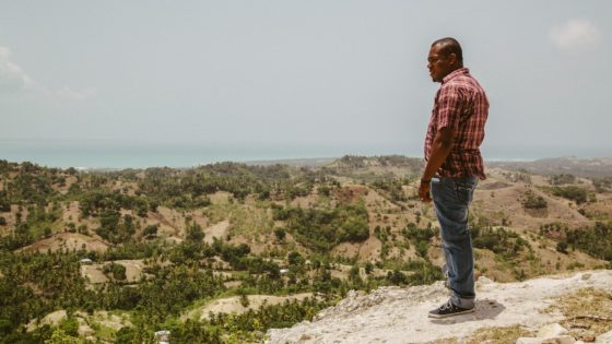 A Haitian man wearing a plaid tan shirt stands on a hillside looking over the valleys and hills below him.