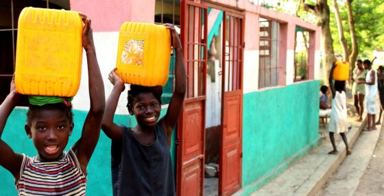Two Haitian children carrying yellow plastic water buckets on their heads. Behind them is a small water supply building with its door open. In the background are more people carrying yellow plastic water buckets on their heads.