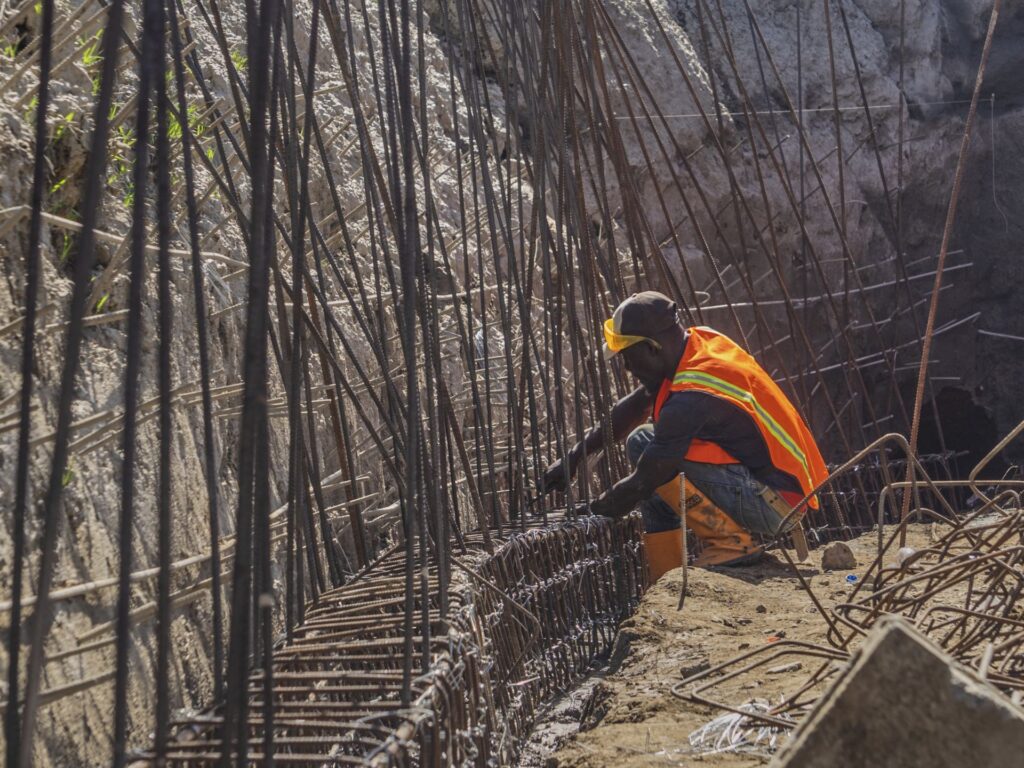 Man wearing hard hat and orange safety vest is tying iron construction rods together.