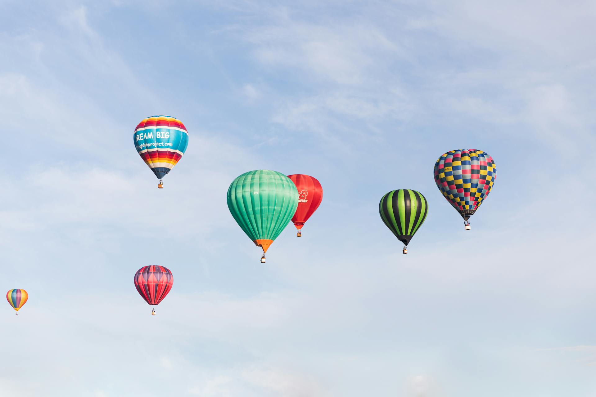 Seven hot air balloon in various colors float in the light blue sky.