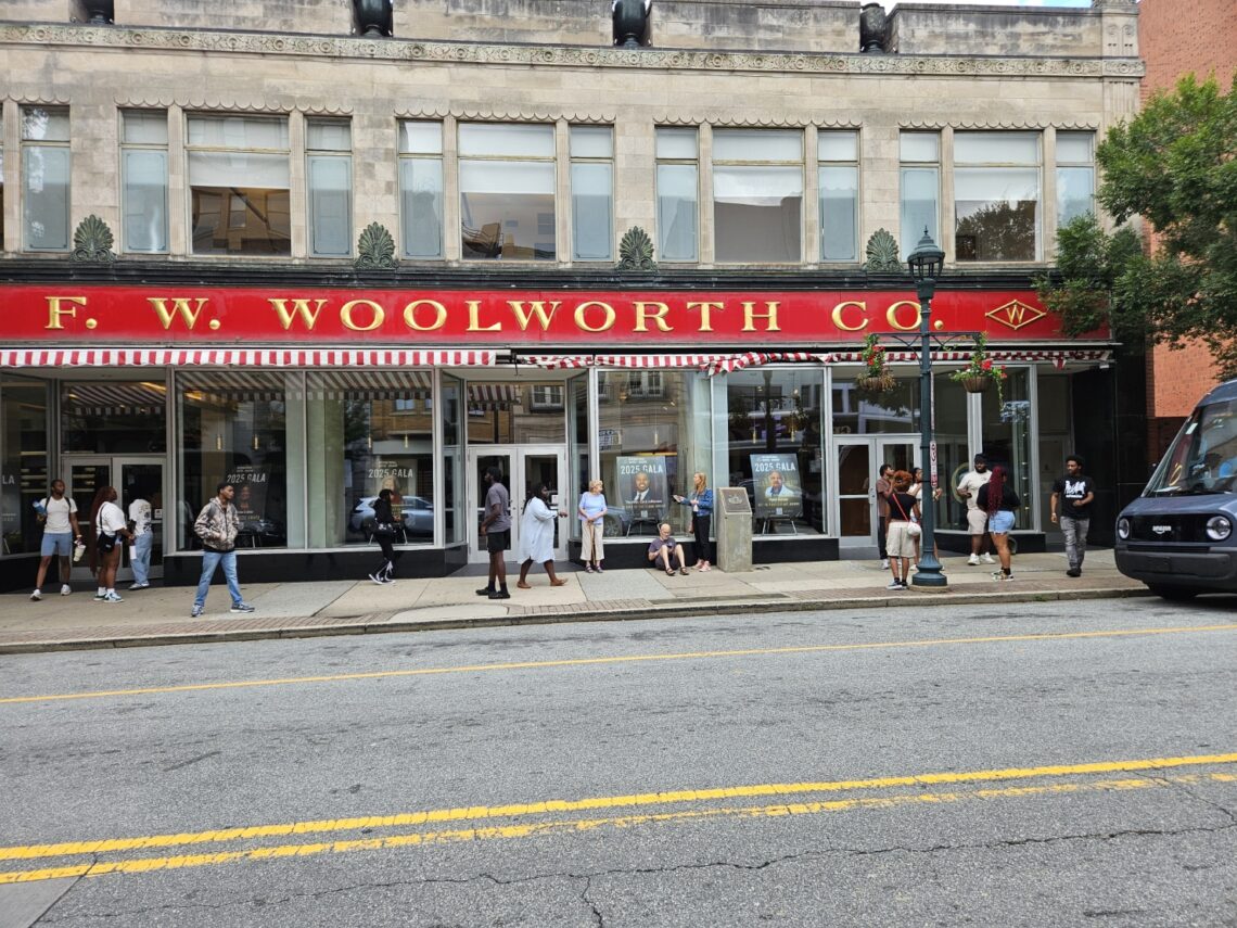 Street view of the F. W. Woolworth building in Greensboro, North Carolina. There are a dozen people standing on the sidewalk in front of the building.