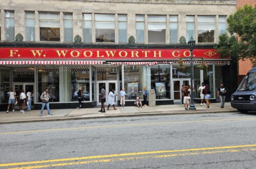 Street view of the F. W. Woolworth building in Greensboro, North Carolina. There are a dozen people standing on the sidewalk in front of the building.