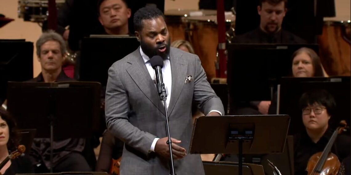 A Black man speaks in front of an orchestra.