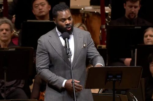 A Black man speaks in front of an orchestra.