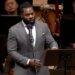 A Black man speaks in front of an orchestra.