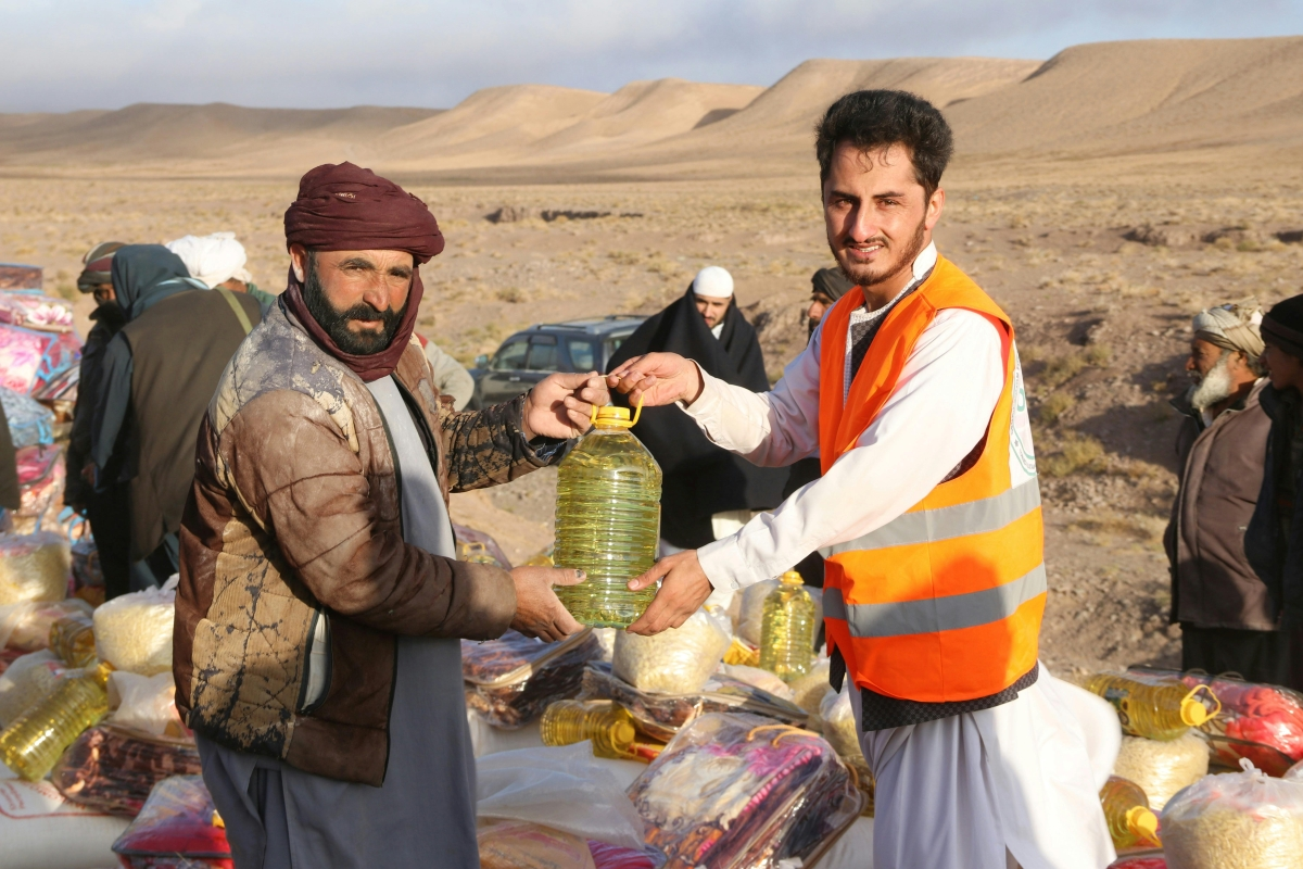 Two men in the high desert of the Near East exchange a water jug. One is wearing an orange vest over a white long-sleeve shirt, and the other is wearing a brown jacket over a long blue shirt or kaftan