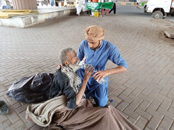 an older man in an old sleeping bag in a public area, leans up from his position to accept water from a younger man helping him drink.