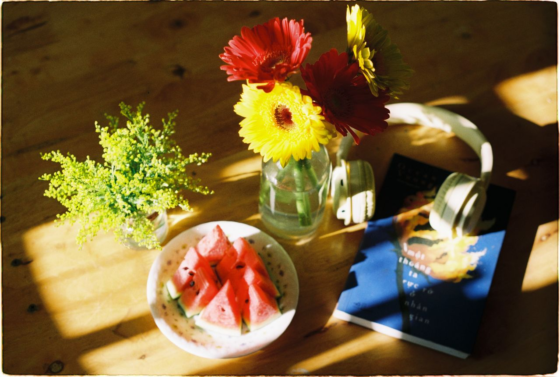 A warm, wooden table with yellow sunlight shining on it at a slant. There is a bunch of yellow-green flowers, a bowl of cut watermelon, a blue book with a pair of white headphones on it, and a bright yellow sunflower, cut short, and put in a glass jar with water.